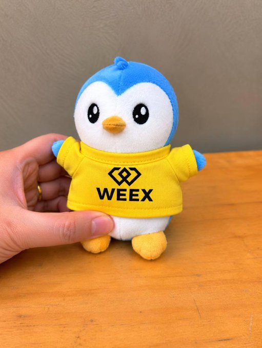 A plush Pudgy Penguin mascot wearing a yellow shirt displaying the "WEEX" logo, being held by a hand. The image ties to the post's announcement of a WeEX × Pudgy Penguins collaboration at Token2049, promoting their upcoming community-driven meme campaign at the event.