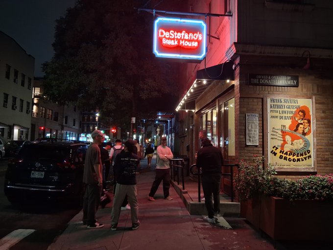 A group gathers on a city sidewalk at night outside DeStefano's Steak House, its iconic red neon sign glowing above as the attendees mingle on the street. The post's mention of an "alpha holder dinner" with the "WEB3 community" fits the lively, after-hours meetup vibe outside this Brooklyn establishment, where they're celebrating "the keys to the city" atmosphere.