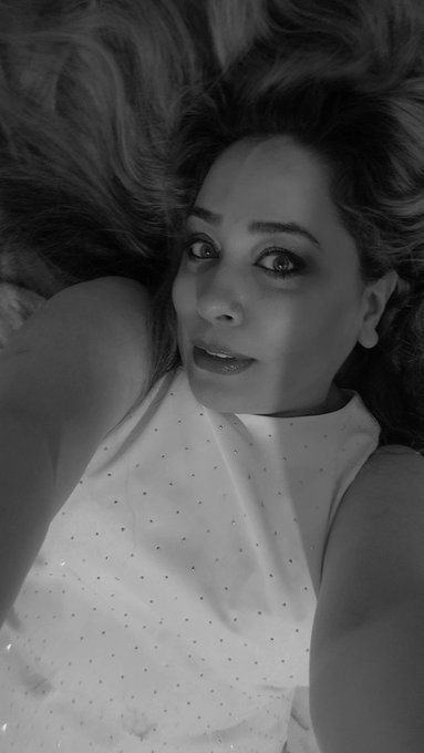 A black and white close-up selfie of a woman with long wavy hair wearing a white sleeveless top with small dotted details, looking at the camera.
