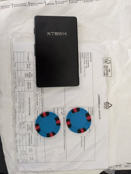 A black 'XTEINK' e-reader sits atop a plastic shipping bag containing a commercial invoice and two blue poker chip-like objects.