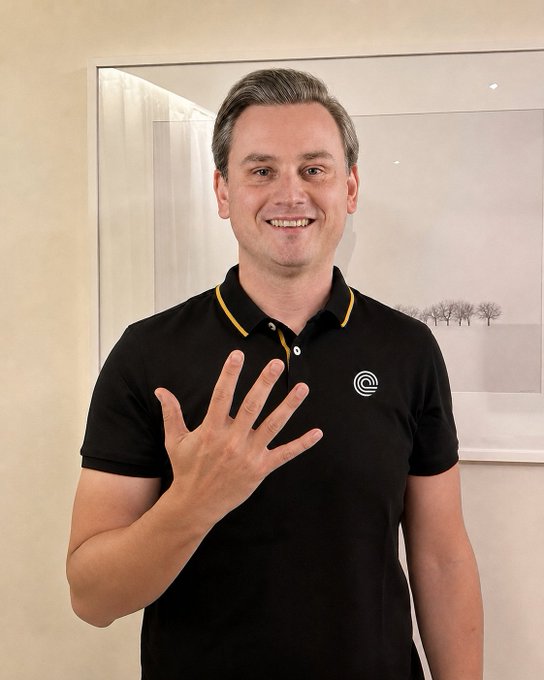 A smiling man in a black polo shirt with a circular logo stands indoors against a wall, holding up his hand with all five fingers extended, symbolizing the "$5 billion" milestone referenced in the post about Ondo Perps reaching that figure in approximately three weeks.