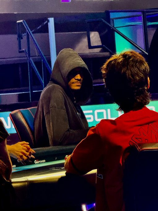 A hooded figure, likely Ansem, sits at what appears to be a poker or gaming table with "GOSPLAYER" branding visible, while another person in a red jacket faces him across the table. The scene suggests an interview or casual meeting setting, fitting the post about Ansem surpassing Nikita Bier in X (Twitter) followers.