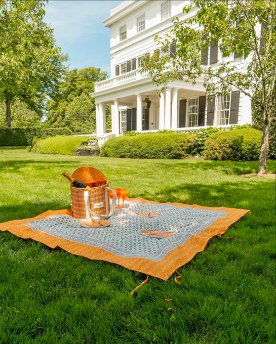 A picnic blanket with a wicker basket, wine bottle, and glasses is spread out on a lush green lawn in front of a large white colonial-style house.