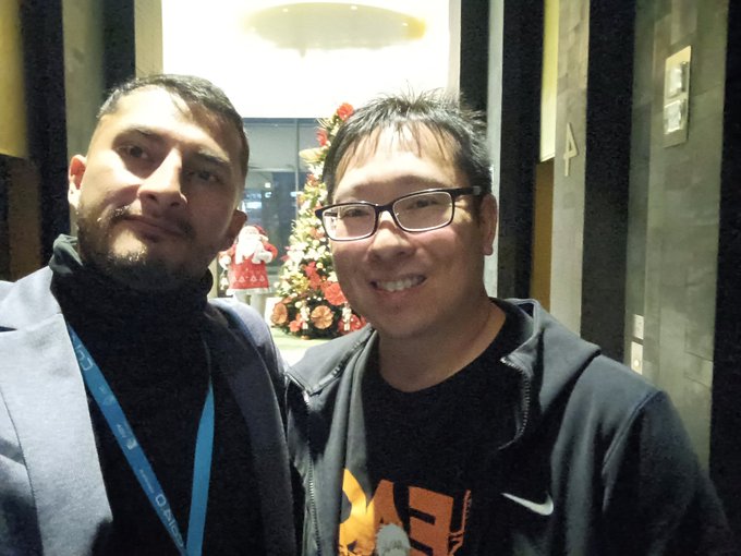 Two men pose for a selfie in a hallway, with a Christmas tree and decorations visible in the background.