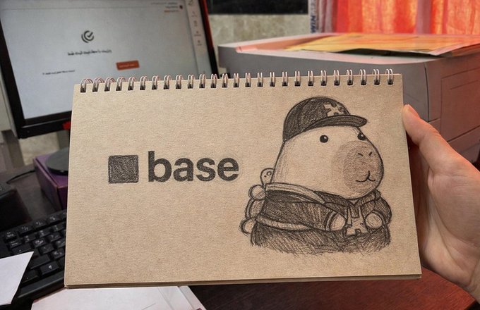 A hand holding a spiral-bound sketchbook with a pencil drawing of a cartoon character wearing a baseball cap and the 'base' logo on the cover, displayed in an office setting.