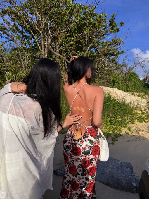 Two women stand from behind on a beach, one wearing a floral skirt and a crisscross-back top with the watermark 'chog', and the other in a white sheer cover-up.
