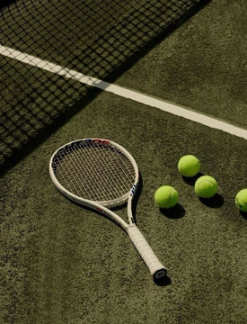 A tennis racket lies on a green tennis court next to three yellow tennis balls, with the net's shadow visible across the court.