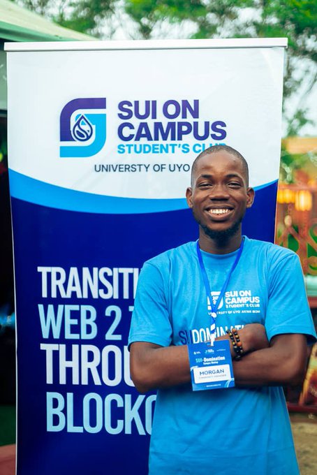 A young man smiling and crossing his arms while wearing a blue 'Sui On Campus Student's Club' t-shirt, standing in front of a banner for the University of Uyo, with text mentioning 'Transition Web 2 To Web 3 Through Blockchain'.