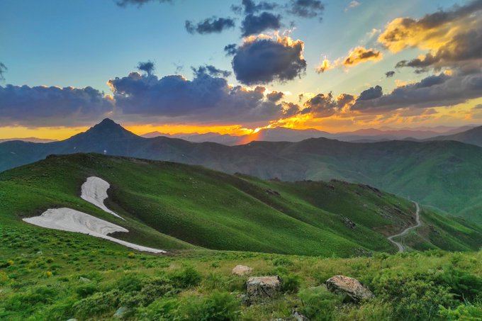 A scenic landscape photograph taken at sunset, showing rolling green hills, a patch of snow on the left, a distant mountain peak, and a colorful sky with dramatic clouds over a winding valley road.
