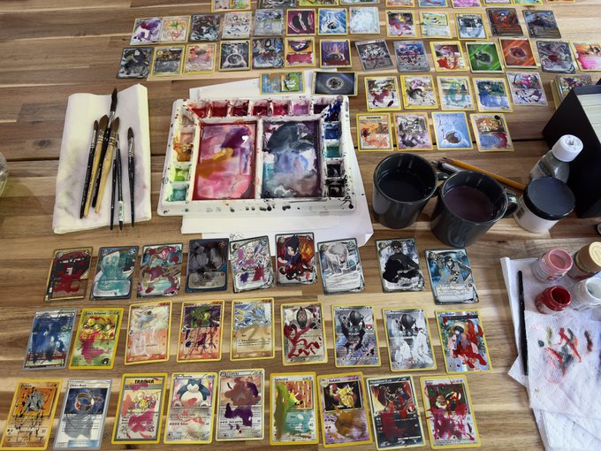 A wooden table is covered with dozens of hand-painted Pokémon cards, including recognizable names like "Magnemite" and "Farfetch'd," alongside a well-used watercolor palette, brushes, and jars of pigment, illustrating the extensive physical craftsmanship behind the digital NFT process.