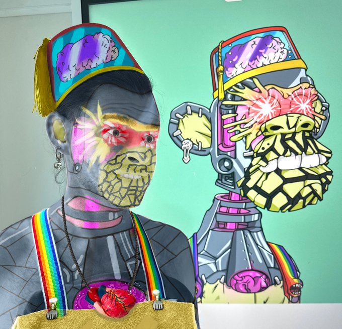A person with elaborate face paint standing next to a colorful cartoon-style drawing of a robot character wearing a hat with brains, set against a mint green wall.