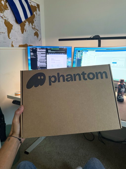 A person holds a brown cardboard box with the "phantom" logo (a ghost face) in front of a dual-monitor desk setup, with a Greek flag and a world map poster on the wall behind.