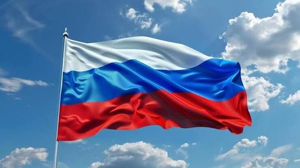 The Russian flag with its white, blue, and red horizontal stripes waves against a bright blue sky with scattered white clouds.