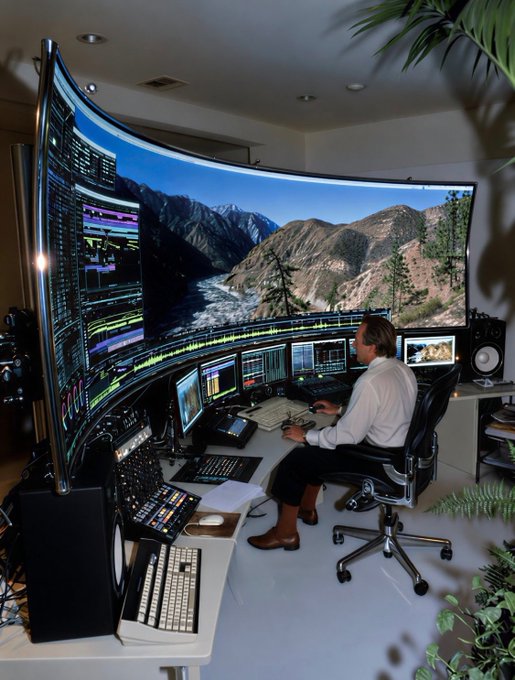 A man working at a sophisticated, curved multi-monitor trading or editing workstation displaying mountain landscapes and data charts.