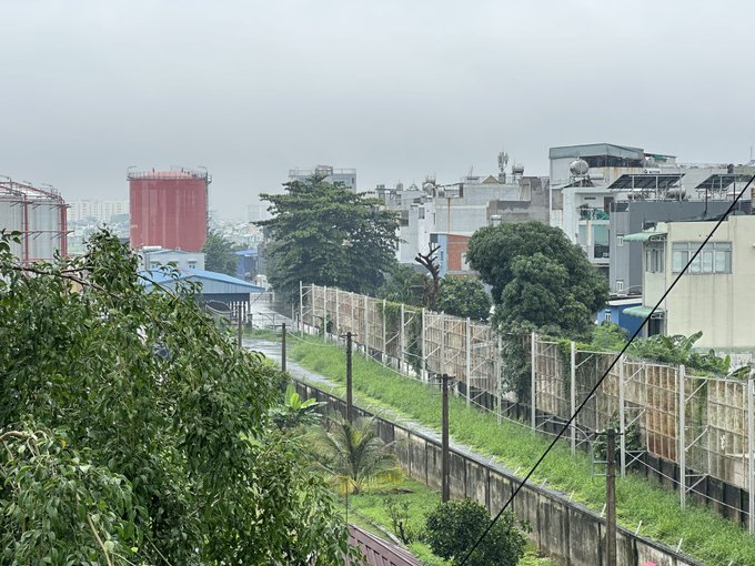 An overcast daytime view of an urban industrial area featuring a grassy embankment with a metal fence, a chain-link fence with billboards, red storage tanks, and residential buildings in the background.