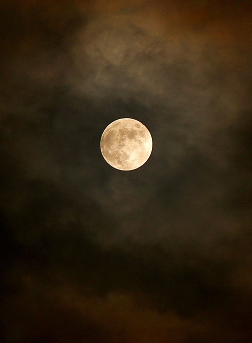 A full moon glowing yellow-orange through dark, wispy clouds in the night sky.