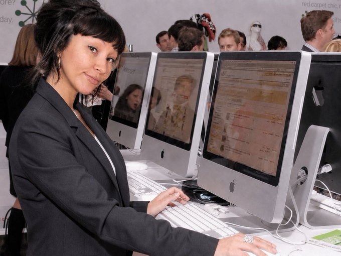 A woman in a black blazer sits at an iMac computer in what appears to be a busy tech event or showcase, with multiple Apple computers lined up on display.