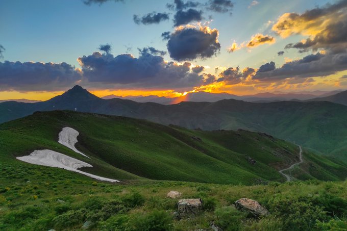 A scenic mountain landscape at sunset with green rolling hills, patches of snow, and dramatic orange and blue clouds across the sky.