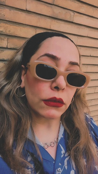 A close-up selfie of a woman wearing small oval sunglasses, red lipstick, hoop earrings, and a blue floral shirt against a brick wall.