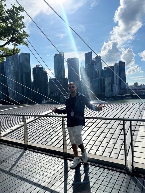A man stands on a bridge with a city skyline in the background, arms outstretched with a confident pose, perfectly capturing the upbeat energy of a "GM" (good morning) post.