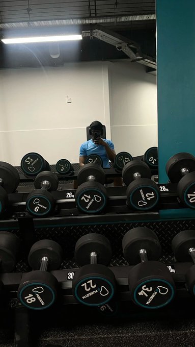 A mirror selfie in a gym showing a person wearing a cap standing behind a rack of dumbbells ranging from 12 to 30 pounds.