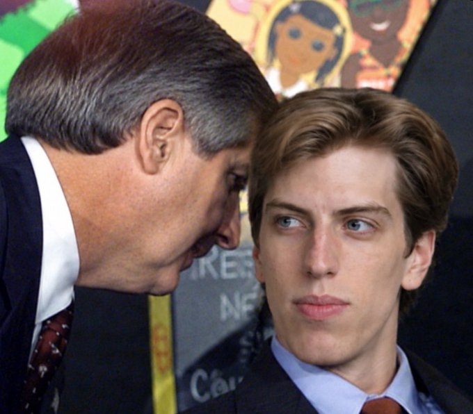A photograph showing a young man with brown hair standing next to an older man with grey hair in a suit, who appears to be whispering to him.