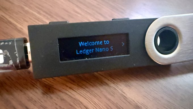 A faded Ledger Nano S hardware wallet displays the welcome message "Welcome to Ledger Nano S" on its dim screen, illustrating the author's warning about screen degradation over time. The barely visible display demonstrates the urgency of transferring funds to a new device before the screen fails completely.
