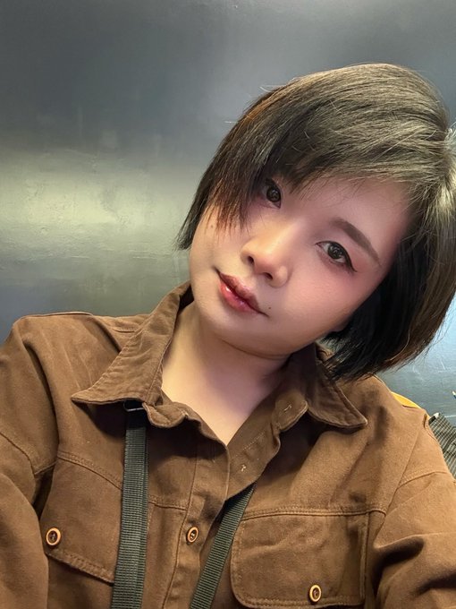 A person with short brown hair wearing a brown corduroy shirt poses for a selfie against a dark background.