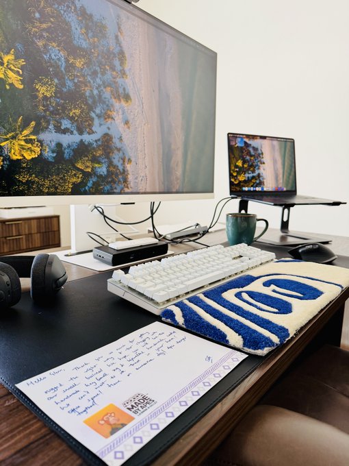 A desk setup with a monitor displaying a forest landscape, a white keyboard, a laptop on a stand, and an envelope with handwritten text addressed to 'Guess'.
