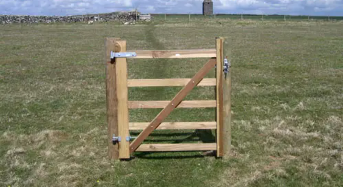 A wooden gate with metal latches stands in a grassy field, with stone walls and a tower visible in the background.
