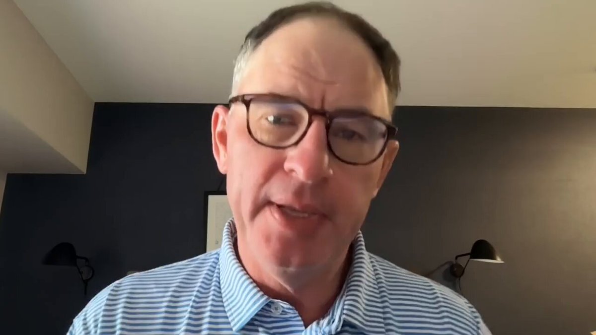The image features a man wearing glasses, standing against a black background with two lamps behind him. He appears to be looking at something off-camera or engaged in an activity that requires his attention. The caption of the image reads "XRP Millionaire Thesis Watch The Full Youtube Video Here: youtu.be/-hv2U1Yxg2o". This suggests that the man might be watching a video related to cryptocurrency, such as discussing XRP or analyzing its value in the market.