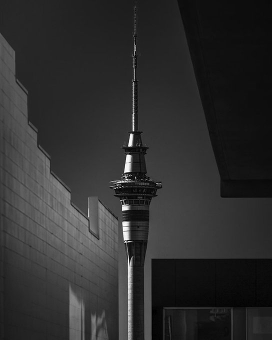 A black and white photograph of a tall communication/broadcast tower rising against a dark sky, framed by modern building architecture in the foreground.