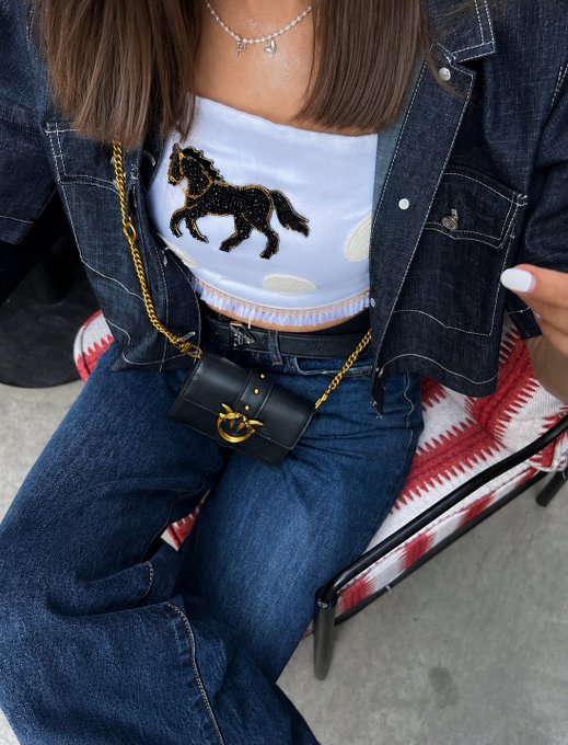 A close-up of a person wearing a light blue top with a black horse design, a dark denim jacket, jeans, and a small black Chanel crossbody bag with a gold chain.