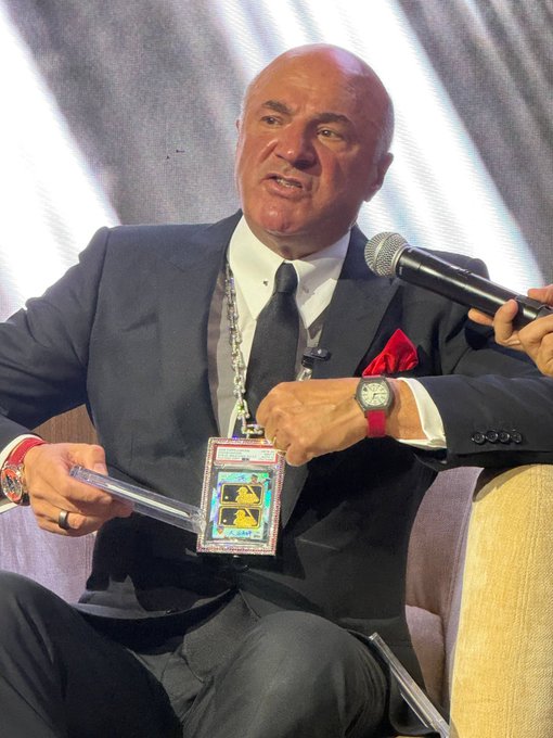 A bald man in a dark suit with a red pocket square speaks into a microphone while wearing a credential lanyard and displaying a red watch.