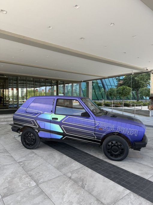 A vintage three-door hatchback car with a custom purple and green cyberpunk wrap featuring the text 'SOLANA', parked in a modern building's driveway.