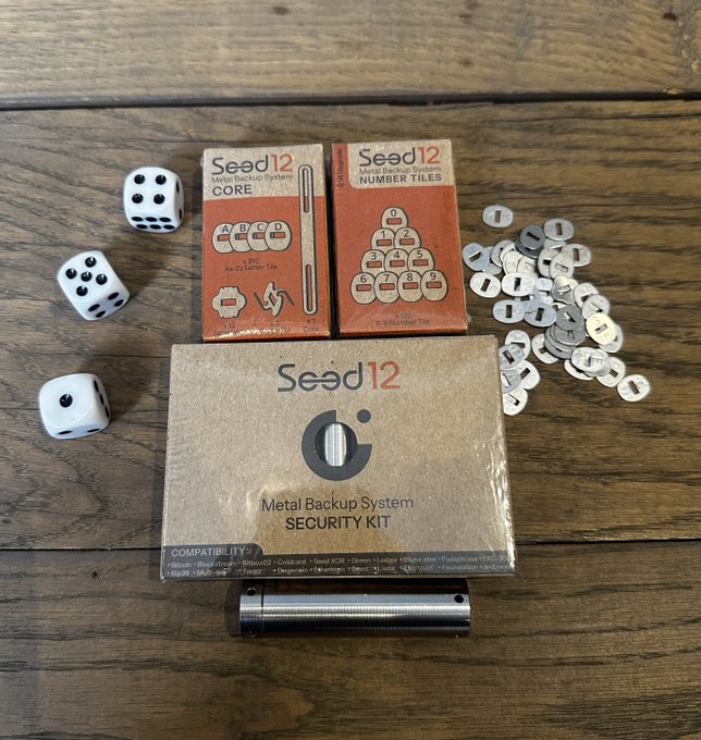 Components of the Seed12 Metal Backup System laid out on a wooden surface, including the Core box, Numbers tiles box, Security Kit box, a metal cylinder, letter tiles, and three dice.