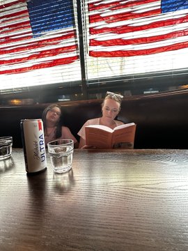 Two women sit at a wooden table in a restaurant, with one reading a large "Big Print" book while a Michelob Ultra can and a glass of water sit on the table in front of an American flag window display. The caption reflects the moment the reader unknowingly picked up a book by author Lawrence Lepard, to her companion's excitement.