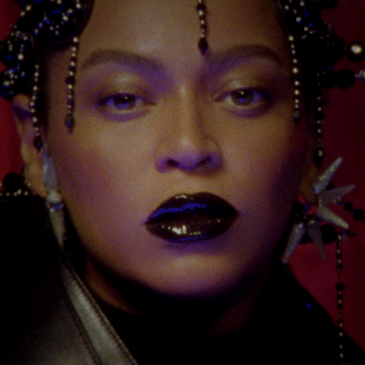 A close-up of a woman with braided hair adorned with beads, dark glossy lipstick, and a star-shaped earring set against a red background, evoking a dramatic and stylized pop aesthetic that visually underscores the sense of a major missed opportunity or loss.