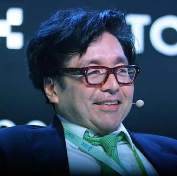 A man with dark-framed glasses and a green tie smiles while wearing a headset microphone at what appears to be a conference or interview setting. The image does not directly relate to the post about ETH price forecasts, as no text, numbers, tickers, or names identifying the person or connection to cryptocurrency are visible.