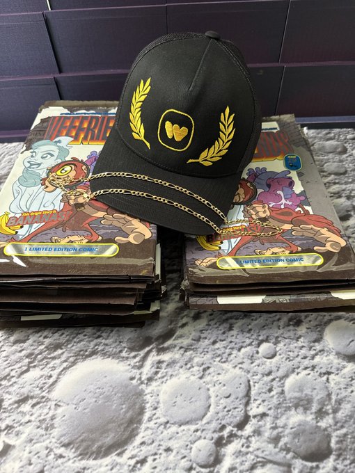 A black baseball cap with gold embroidery showing two hearts flanked by laurel wreaths, displayed on comic books with chains, sitting on a moon-surface textured background.