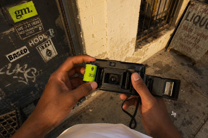 A pair of hands loads a roll of "gm." 300 film into an open camera, preparing to capture "final snaps" on a graffiti-covered city street at sunset. Visible stickers reading "BASED ETH" and "HODL" decorate the doorway beside them, tying into the crypto-themed post about FCFS access to the @OnChainHoodies drop.