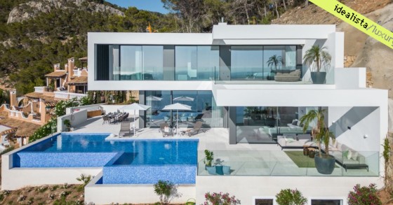 An aerial view of a modern white luxury villa with a large swimming pool, set against a hillside with greenery, sourced from idealista/luxury.