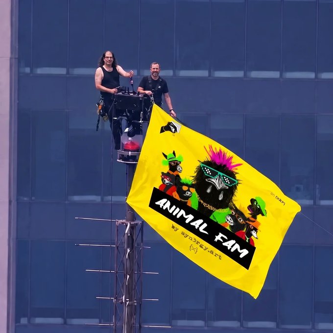 Two men on a tall metal scaffolding/tower holding a bright yellow 'Animal Fam' flag featuring cartoon animal illustrations, with the Instagram handle @graxxkey.art visible.
