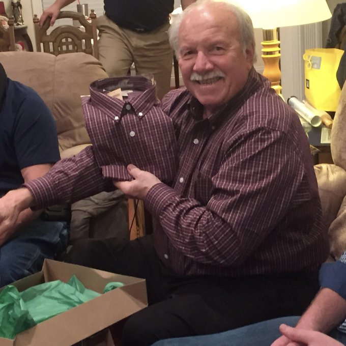 An older man with a mustache sitting on a couch, smiling while holding up a folded purple striped button-down shirt next to an open gift box with green tissue paper.