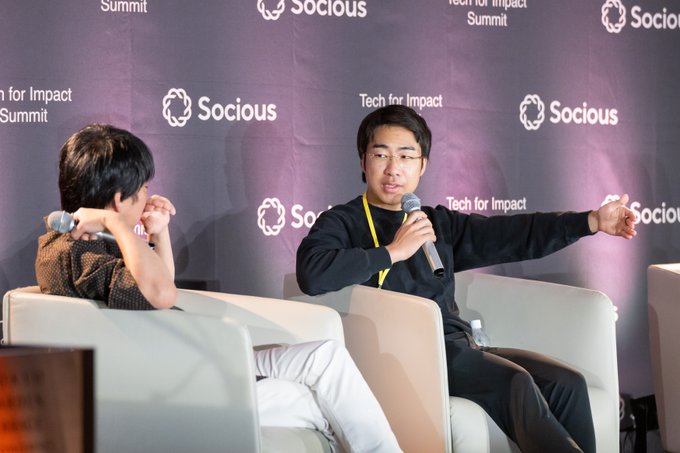 Two men sitting on chairs at the 'Socious Tech for Impact Summit', with the man on the right speaking into a microphone while gesturing.