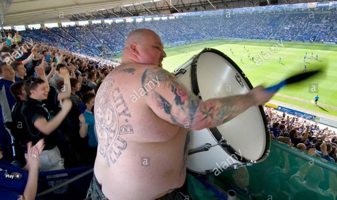 A shirtless, tattooed fan bangs a large bass drum in the stands of a crowded soccer stadium.