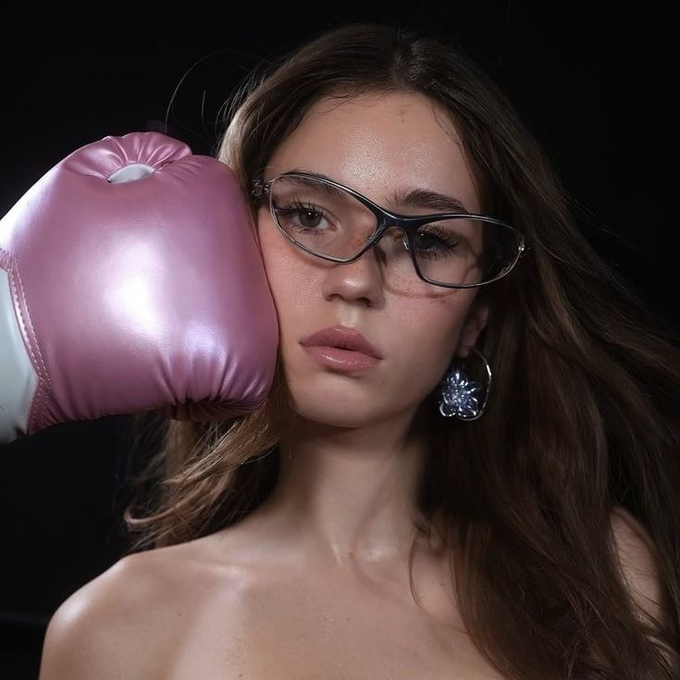 A glamorous portrait of a woman wearing glasses and a pink boxing glove posed against her cheek, juxtaposing the fierce, predatory image of an attacker with news of a $75 million exploit of the Tectonic protocol on the Cronos network, symbolizing the relentless "boxing" of crypto hackers against DeFi projects.