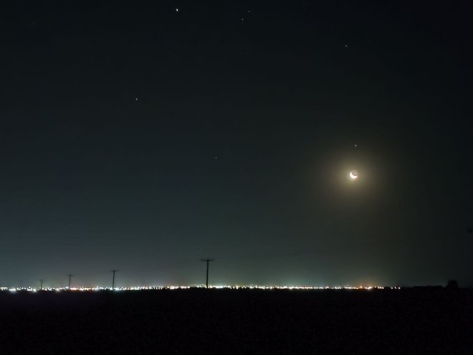 A nighttime photograph showing a crescent moon shining in a starry sky above a distant line of city lights along the horizon.