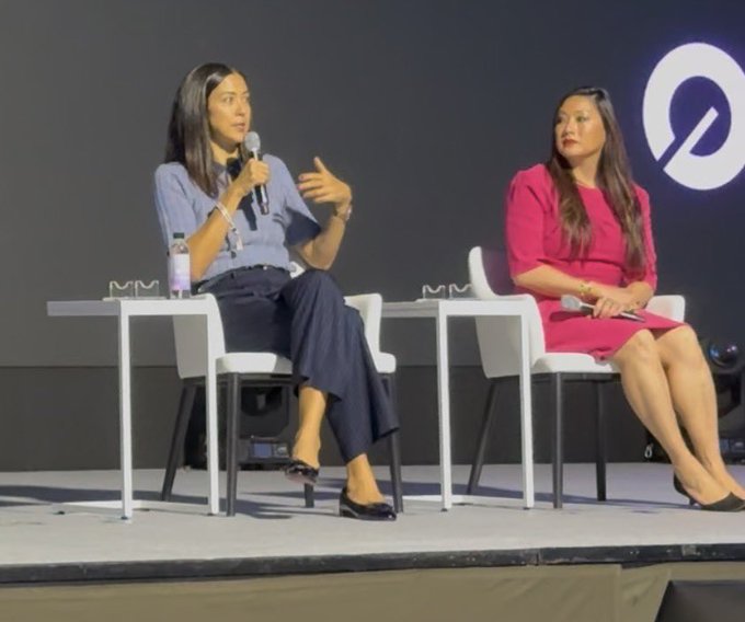 Two women sit on stage at Korea Blockchain Week, with one speaking into it while holding a microphone, engaged in a panel discussion about the next steps for tokenization. The stage features a dark backdrop with a circular logo, and the speakers are seated in white chairs with a small table between them holding water bottles.