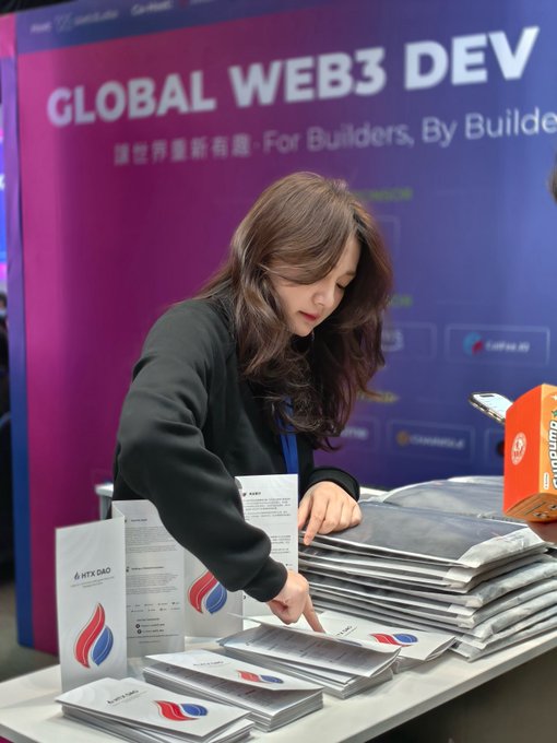 The image features a woman standing at a table, surrounded by several brochures or flyers. She appears to be pointing at one of them while looking down at another flyer. The background shows a purple wall with a logo for Global Web3 Dev on it. There is also an orange box placed near the right side of the scene.